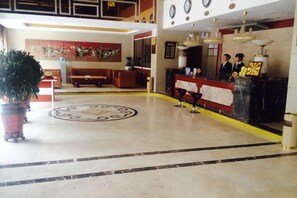 A hotel lobby with a reception desk, a large potted plant, a circular emblem on the floor, and a seating area. - Wulate Hotel (Urad Front Banner)