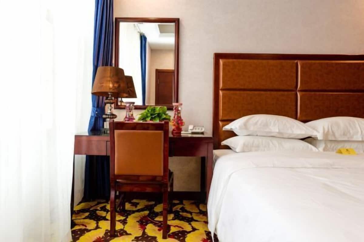 A hotel room with a bed, a desk with a lamp, a chair, a mirror, and a patterned carpet.