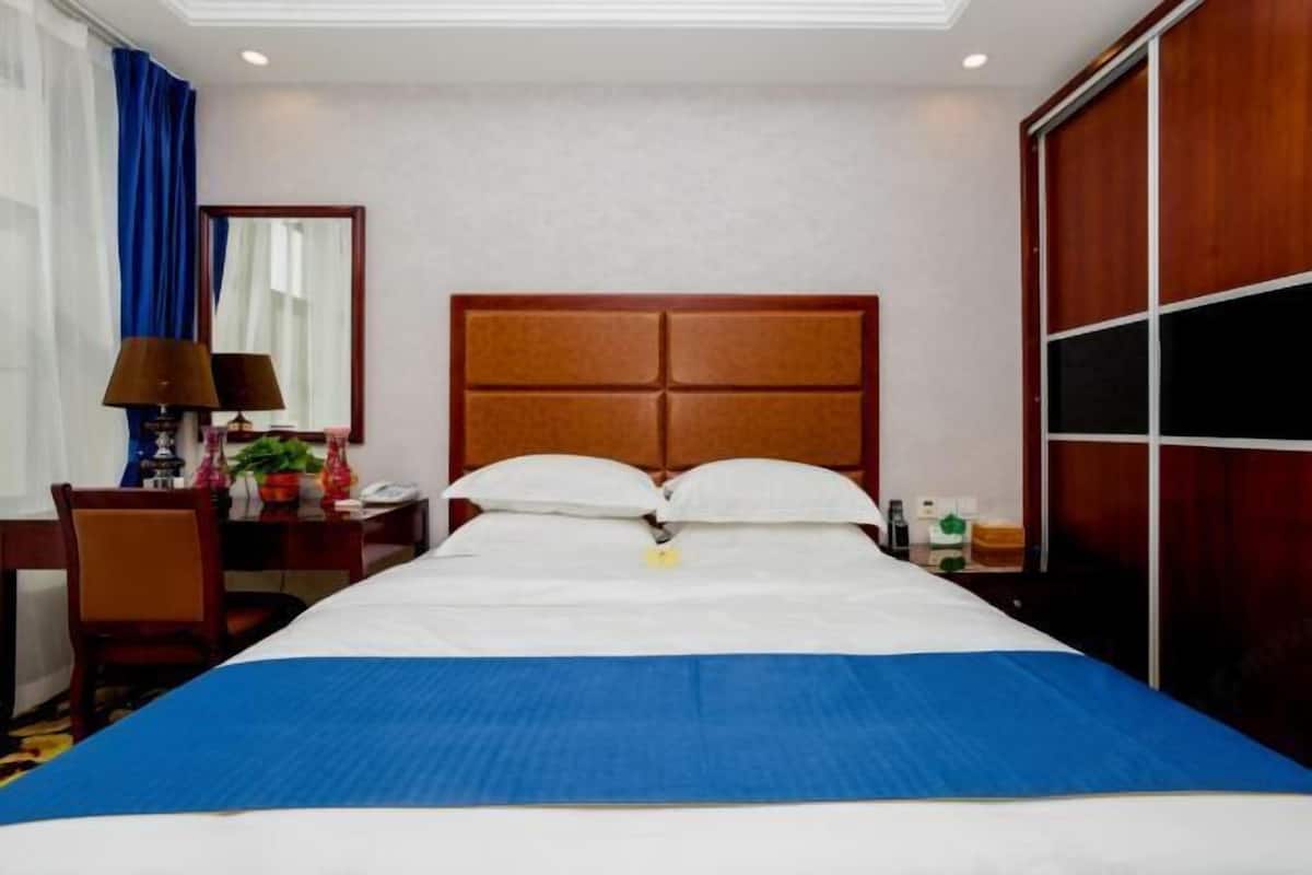 A hotel room with a large bed, a desk with a lamp, a chair, a mirror, and a wardrobe.