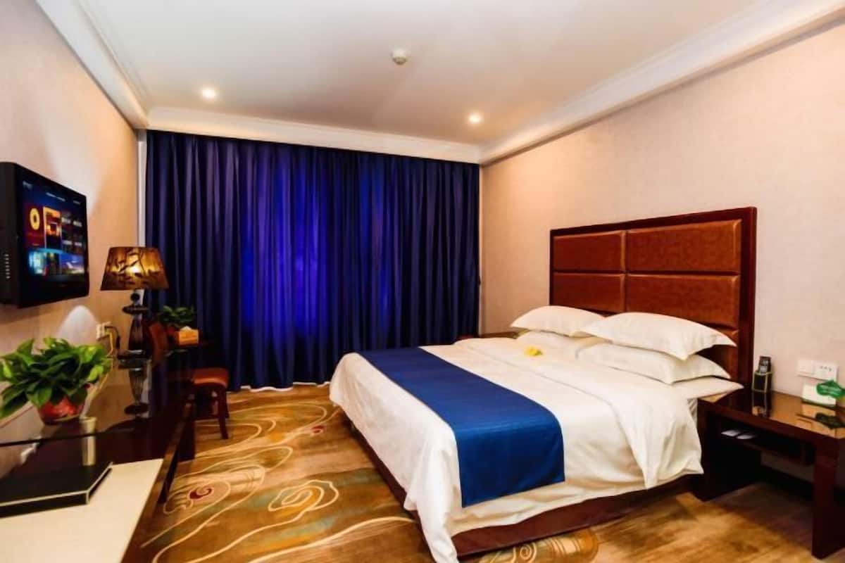 A hotel room with a large bed, a flat-screen TV, a desk with a lamp, and a window with curtains.