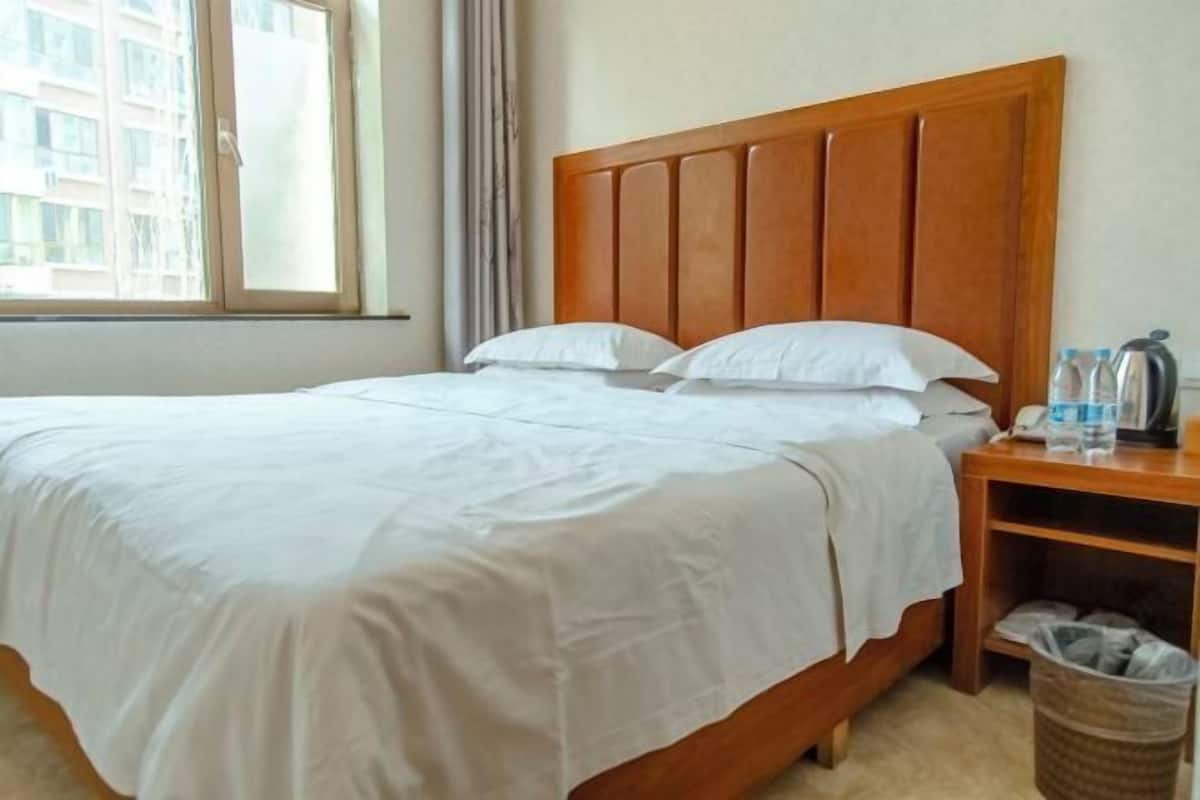 A hotel room with a wooden headboard, a bed with white bedding, a nightstand with a phone and water bottles, and a view of buildings through a window.