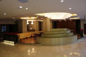 Un vestíbulo de hotel con un mostrador de recepción curvo, una escalera y un área de descanso con mesas y sillas.