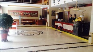 A hotel lobby with a reception desk, a large potted plant, a circular emblem on the floor, and a seating area with a sofa and a bench. - Wulate Hotel (Urad Front Banner)