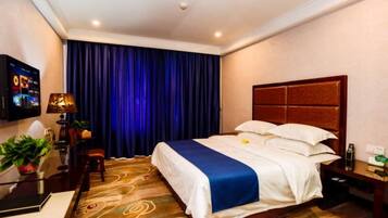 A hotel room with a large bed, a flat-screen TV, a desk with a lamp, and a potted plant.