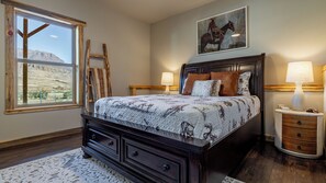 A bedroom with a large bed, wooden headboard, two bedside tables with lamps, a window with a view of a mountain, and a painting of a cowboy on a horse.