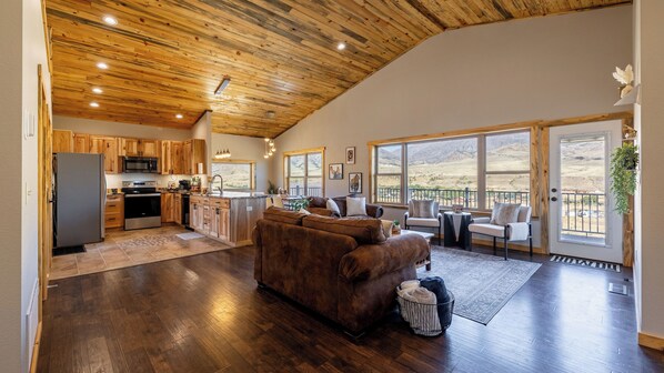 A spacious living area with a large brown leather sofa, a kitchen with wooden cabinets, and a view of the mountains through large windows.