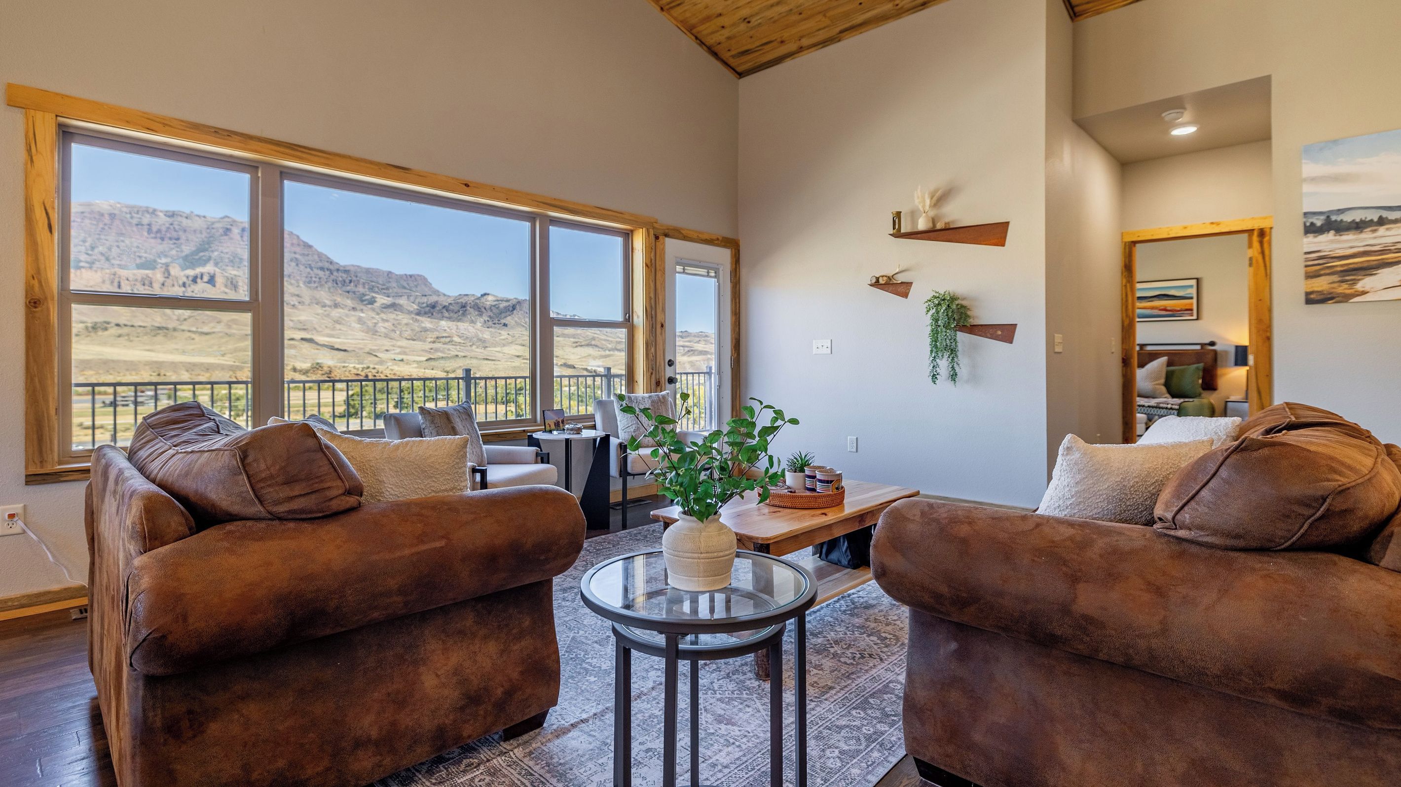 Un salon spacieux avec deux canapés marron, une table basse en verre et une vue sur un paysage montagneux grâce à de grandes fenêtres.