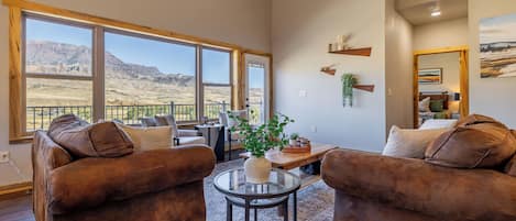 Un salon spacieux avec deux canapés marron, une table basse en verre et une vue sur un paysage montagneux grâce à de grandes fenêtres.