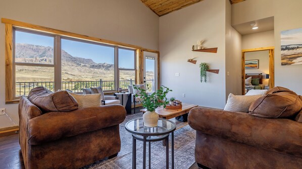 A spacious living room with two brown sofas, a glass coffee table, and a view of a mountainous landscape through large windows.