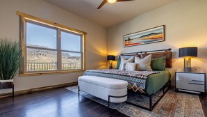 A bedroom with a large window, a bed with a patterned bedspread, two bedside lamps, a nightstand, and a painting on the wall. - Soak in Panoramic Views • Wapiti Cabin, Dogs OK (Cody)