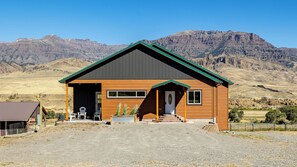 A two-story wooden cabin with a green roof, a front porch, and a gravel driveway.