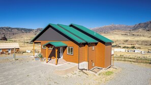 A wooden cabin with a green roof, a porch, and a view of mountains in the background.