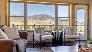 A room with a view of a mountainous landscape through large windows, furnished with a sofa, two armchairs, and a small table.