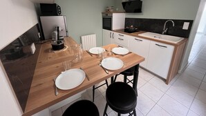 A compact kitchen with a wooden table set for four, a black stool, a black countertop, a white refrigerator, a black oven, a black stove, a black sink, and a black cabinet. - Maison du Colombier (Essarts-en-Bocage)