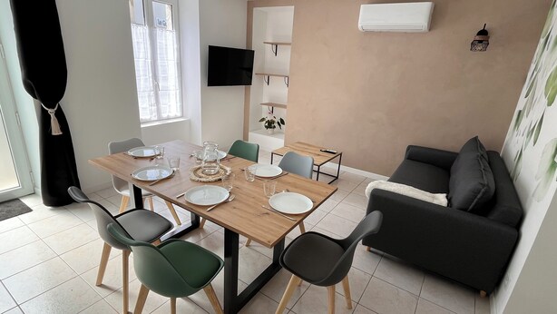 A modern living room with a dining area, featuring a wooden dining table, green and black chairs, a black sofa, and a TV mounted on the wall. - Maison du Colombier (Essarts-en-Bocage)