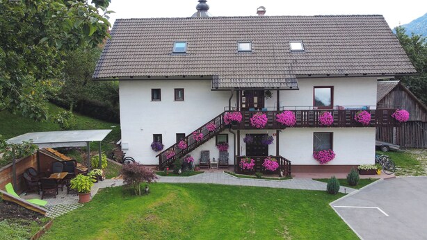 Une maison à deux étages dotée d’un balcon orné de fleurs roses, d’un petit jardin et d’une terrasse en bois.