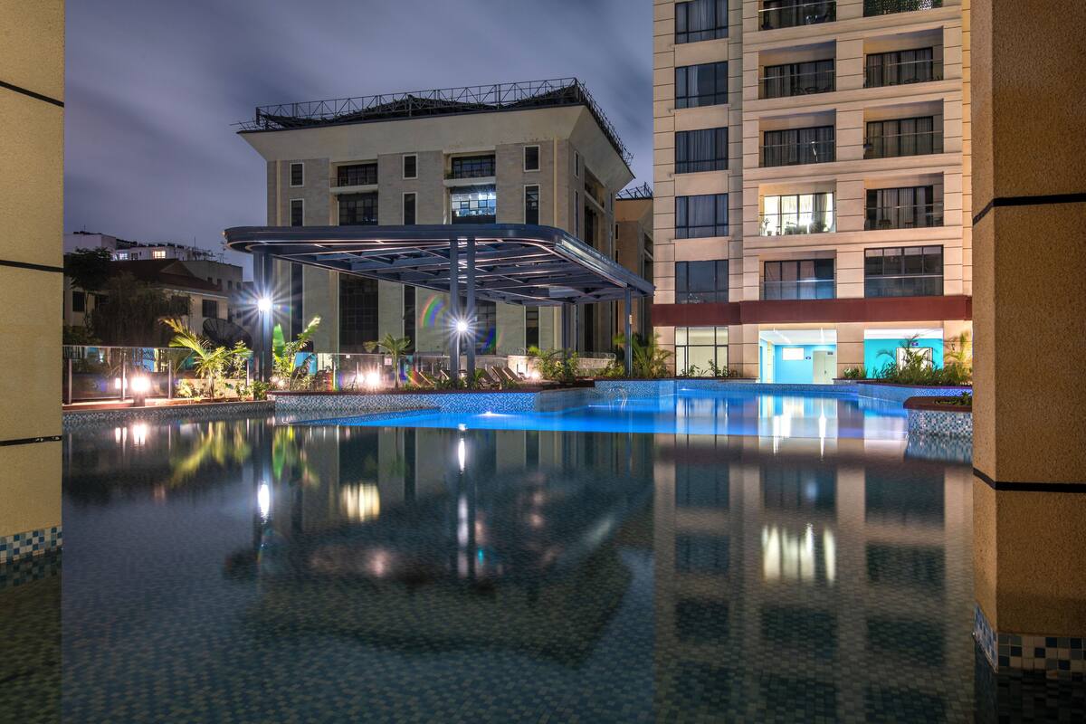 Zona de piscina de hotel con área de estar cubierta, un estanque reflectante y un edificio de varios pisos con balcones.