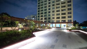 A spacious outdoor area with a tiled floor, surrounded by greenery and a tall building in the background. - Alba Garden (Nairobi)