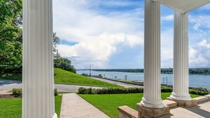 A covered outdoor area with white columns, a grassy lawn, and a view of a body of water. - The Estate at Prospect Park (Davenport)