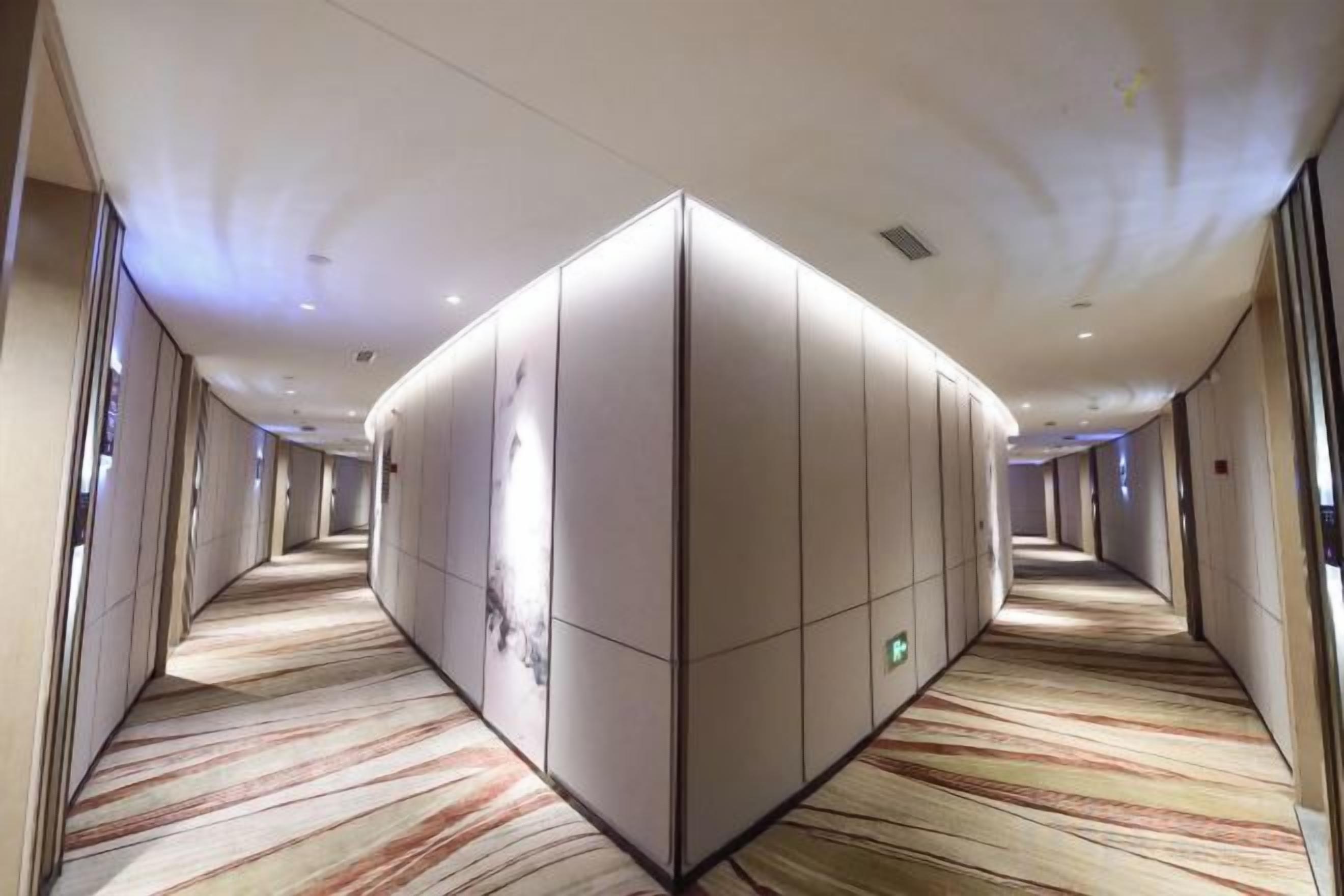 A hotel corridor with a carpeted floor, a central wall with a geometric design, and doors on either side.