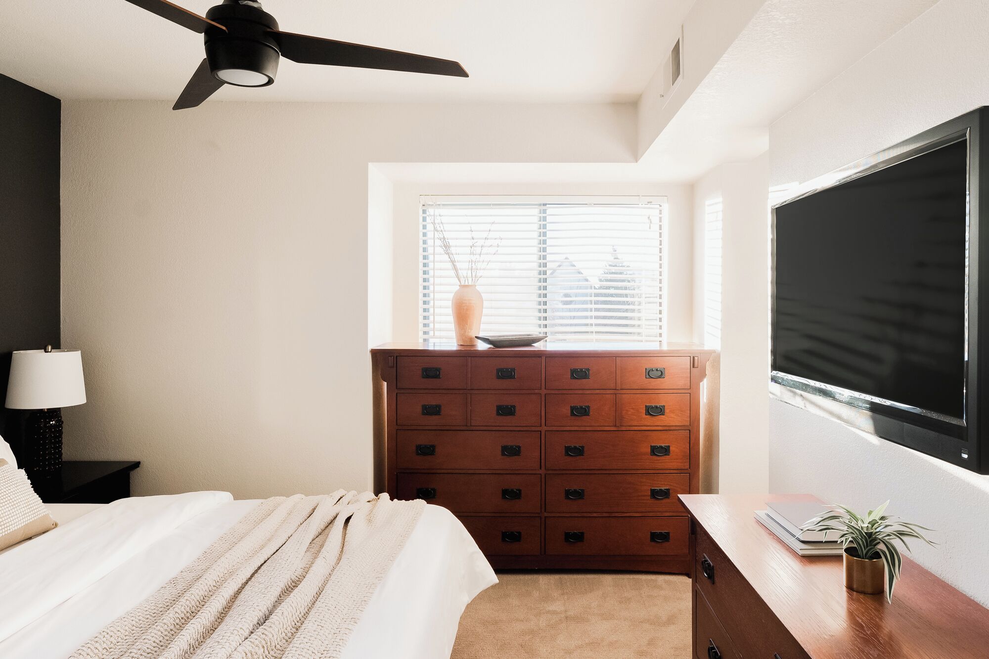 Ein Schlafzimmer mit einem Bett, einer Kommode, einem Schreibtisch, einem Deckenventilator und einem Fenster mit Jalousien.