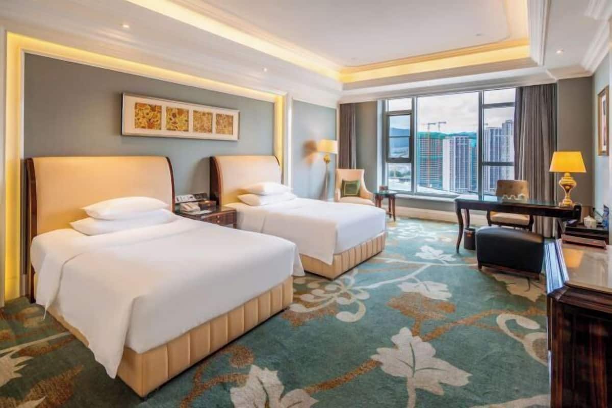 Habitación de hotel con dos camas, un escritorio, una silla y un ventanal con vistas a la ciudad.