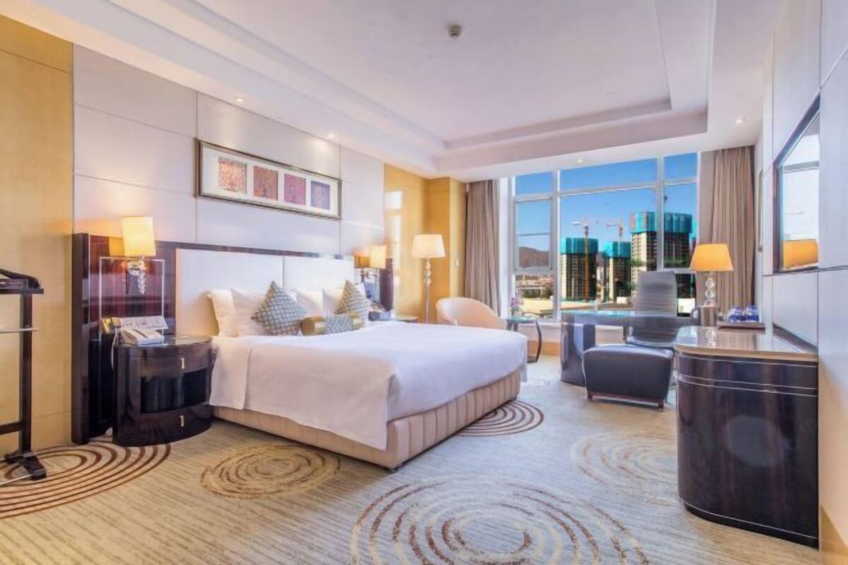 Habitación de hotel con cama, mesitas de noche, escritorio, silla y vistas a la ciudad.