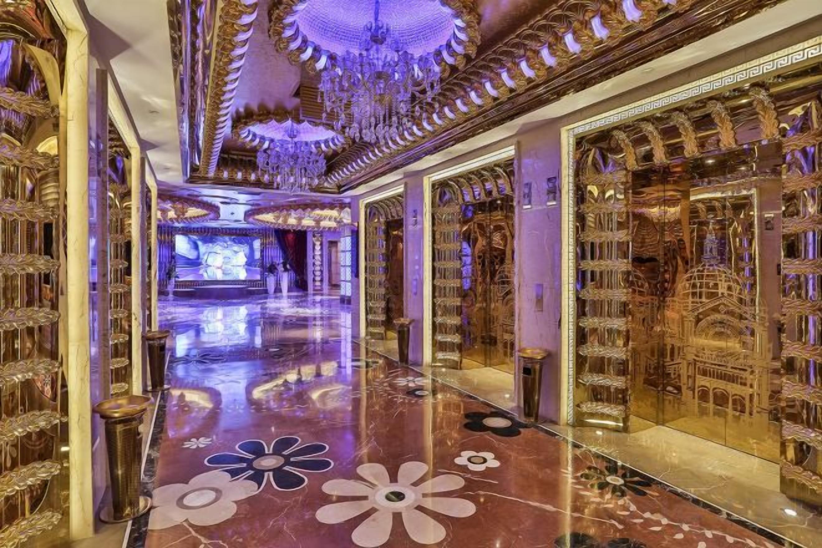 A luxurious hallway with ornate gold decorations, a reflective floor, and a grand chandelier.