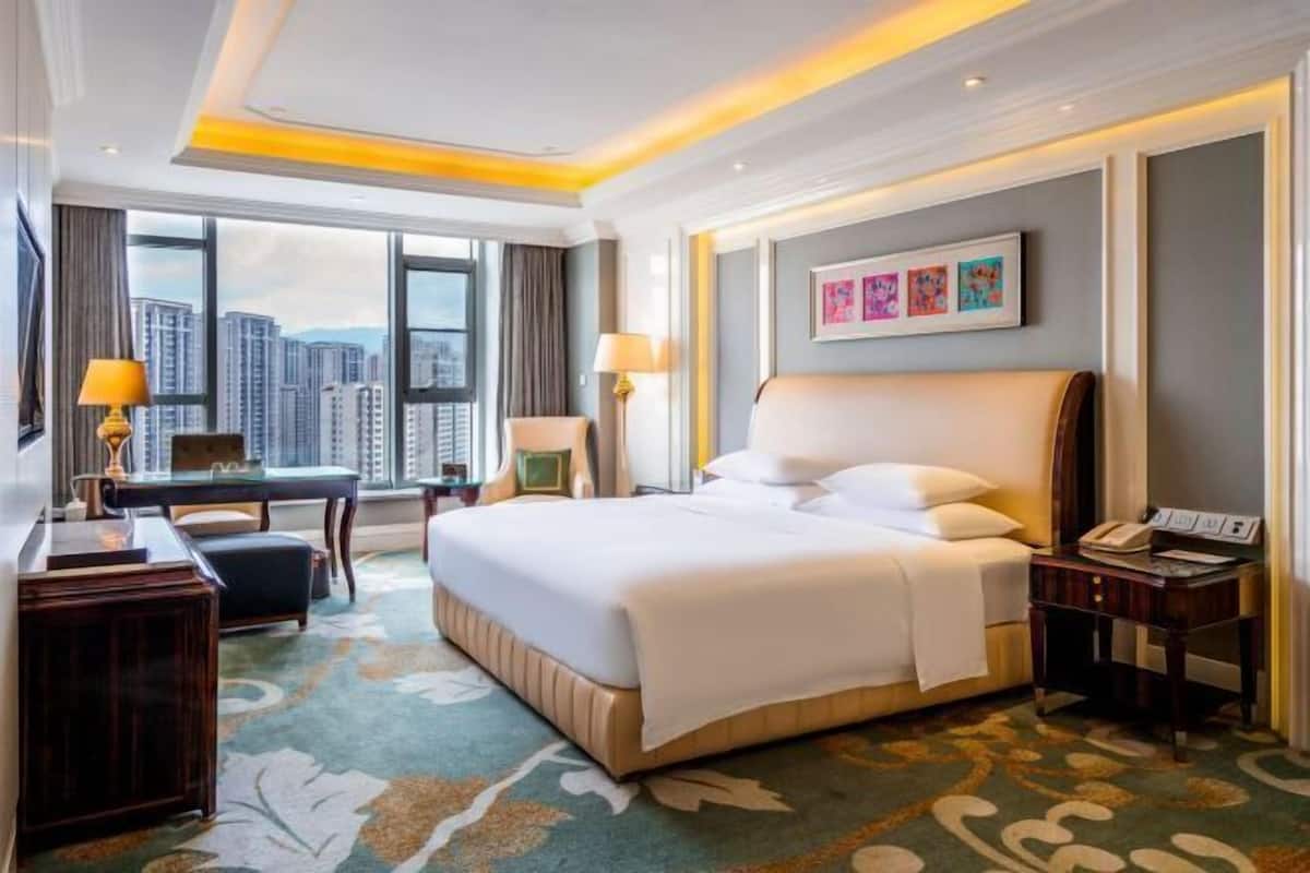 Habitación de hotel con una cama grande, un escritorio, una silla, una lámpara y vistas a la ciudad.