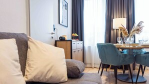 Moderni hotellihuone, jossa on harmaa sohva, puinen sivupöytä lampun kanssa, pieni työpöytä maljakolla ja laseilla sekä suuri ikkuna, josta avautuu näkymä ulos.