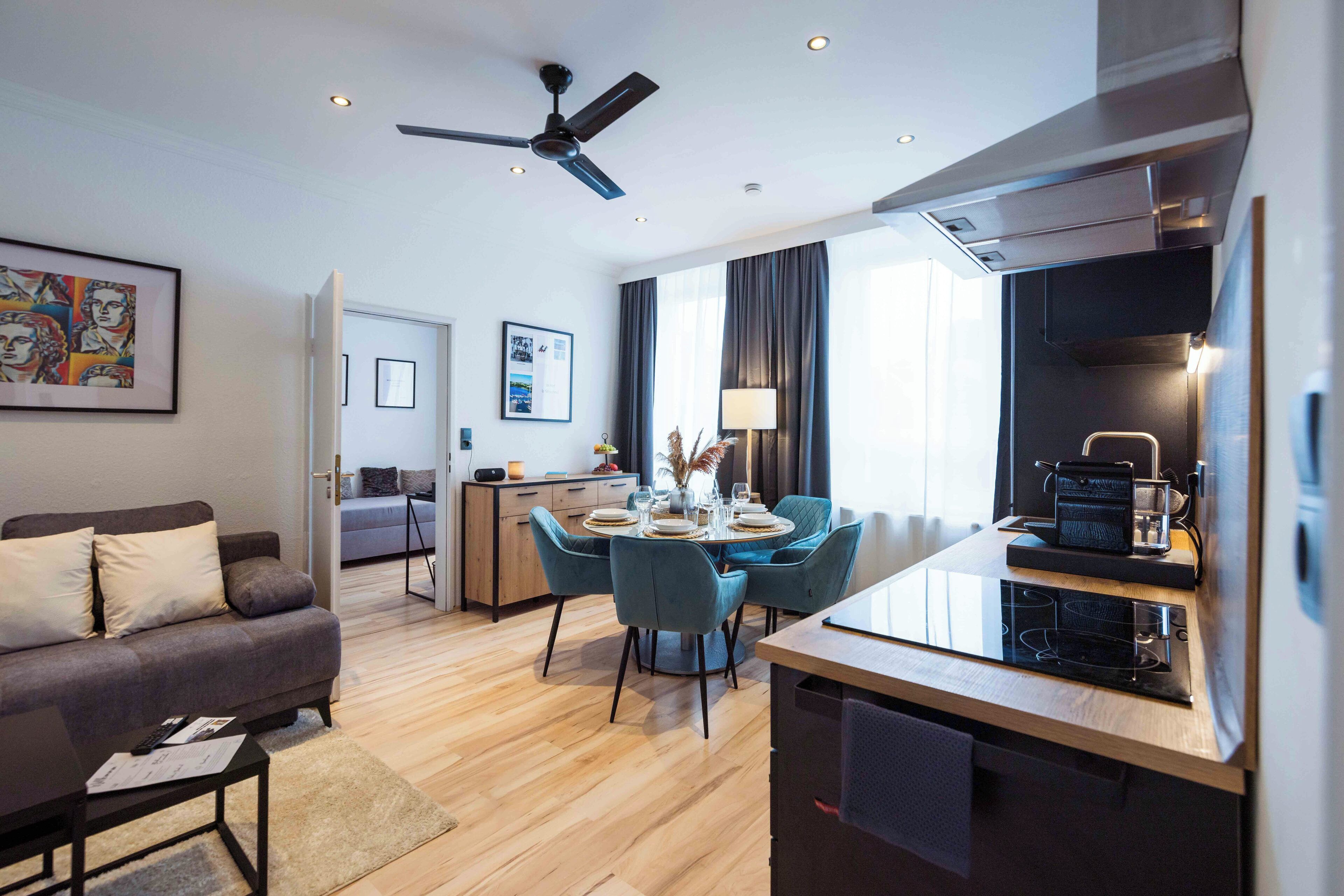 A modern living room with a sofa, a dining table with chairs, a ceiling fan, and a kitchen area with a stove.