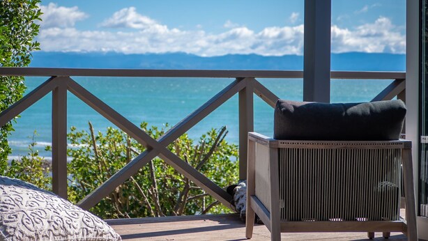 Eine Holzterrasse mit Blick auf den Ozean und die Berge. Ein Sessel mit Kissen und ein kleiner Tisch sind vorhanden.