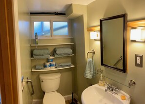 Bathroom features a toilet, sink, and glass shelf with towels, plus a mirror and small window.