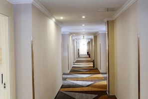 Corredor de hotel comprido e estreito, com carpete estampado, portas em ambos os lados e teto de cor clara.
