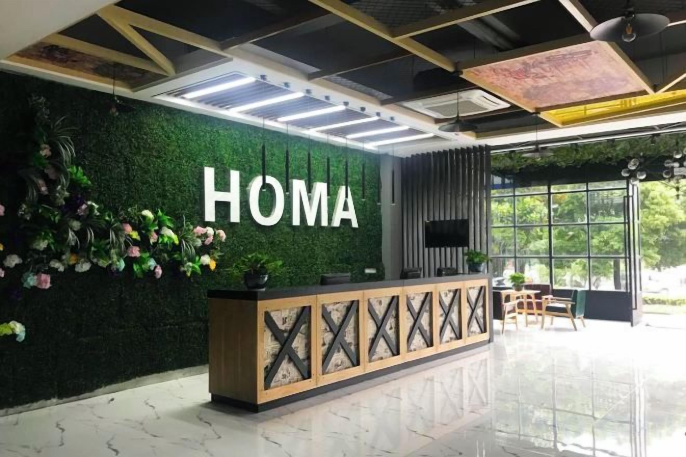 A reception desk with a 'HOMA' sign, a green wall with plants, and a large window providing a view of the outdoors.