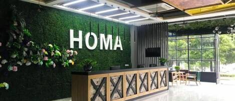 Un mostrador de recepción con un letrero que dice 'HOMA', una pared verde con plantas y un amplio ventanal que ofrece vistas al exterior.