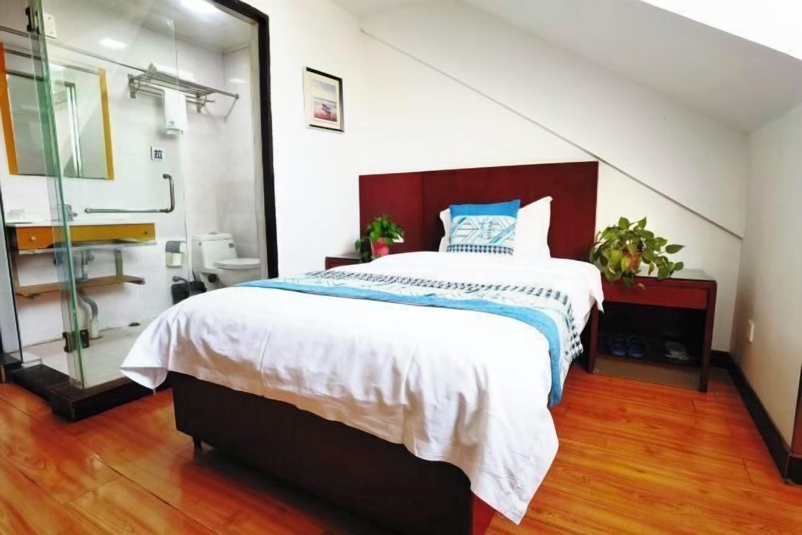 A bedroom with a bed, a nightstand, a potted plant, and a bathroom with a glass shower enclosure.
