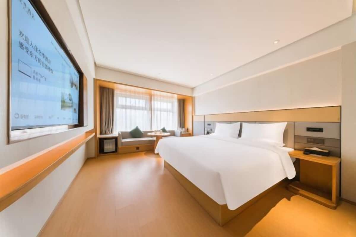 A hotel room with a large bed, a bedside table, a small seating area, and a large window.