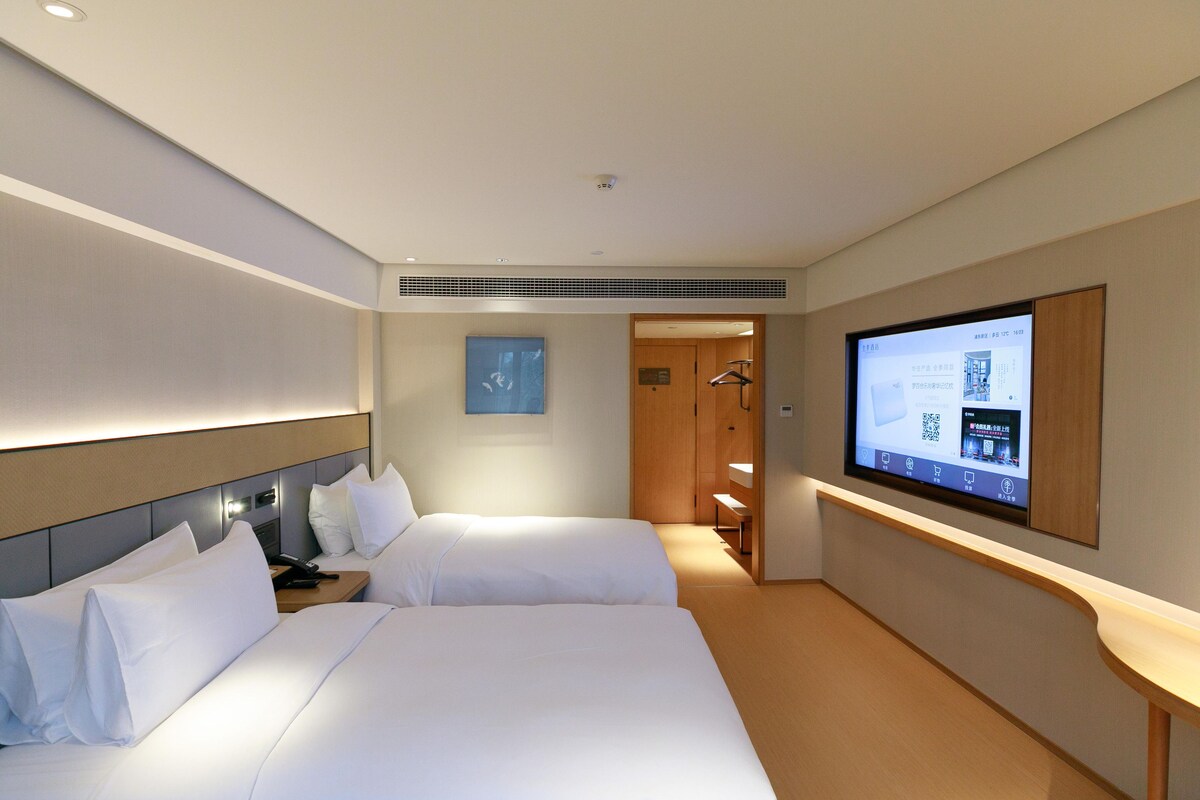 Une chambre d’hôtel avec deux lits, un téléviseur fixé au mur, une porte en bois et un banc.