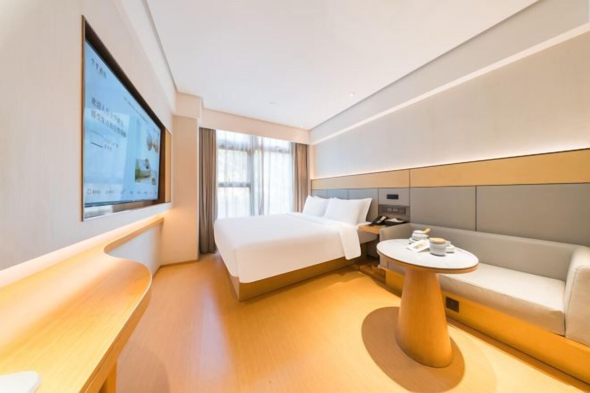 A modern hotel room with a bed, a sofa, a small table, and a TV mounted on the wall.