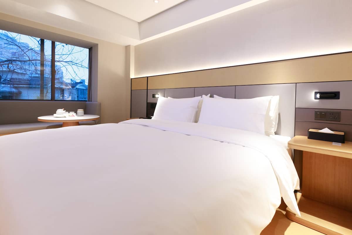A modern hotel room with a large bed, bedside tables, a window with a view, and a control panel on the wall.