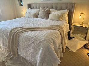 A neatly made bed with a quilted white comforter, a knitted throw, and several pillows. There is a bedside table with a lamp and a mirror. - Kid friendly House on the park (Burnaby)
