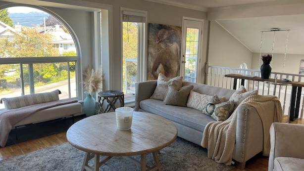 A cozy living room with a large window, a round wooden table, a sofa with multiple cushions, and a painting on the wall.