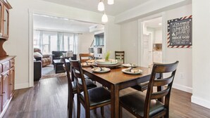 A dining area with a wooden table set for four, surrounded by dark chairs. A chandelier hangs above, and a sign with patriotic text is on the wall. - Pet-Friendly Home with Fenced Yard, Monthly stays (Virginia Beach)