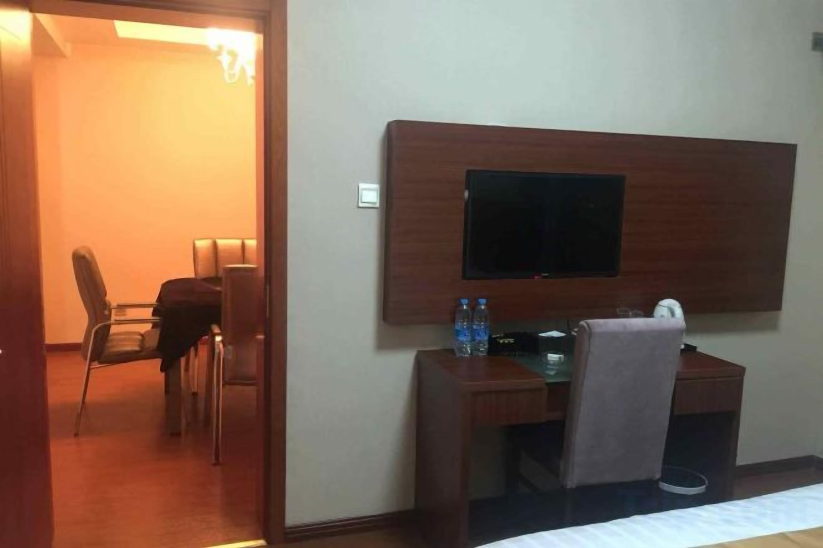 A hotel room with a bed, a desk with a chair, a television mounted on the wall, and a small table with a bottle and a cup.