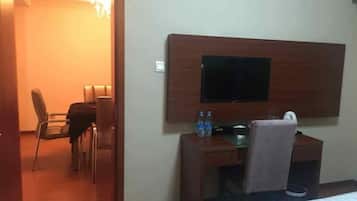 A hotel room with a bed, a desk with a chair, a television mounted on the wall, and a small table with a bottle and a cup.