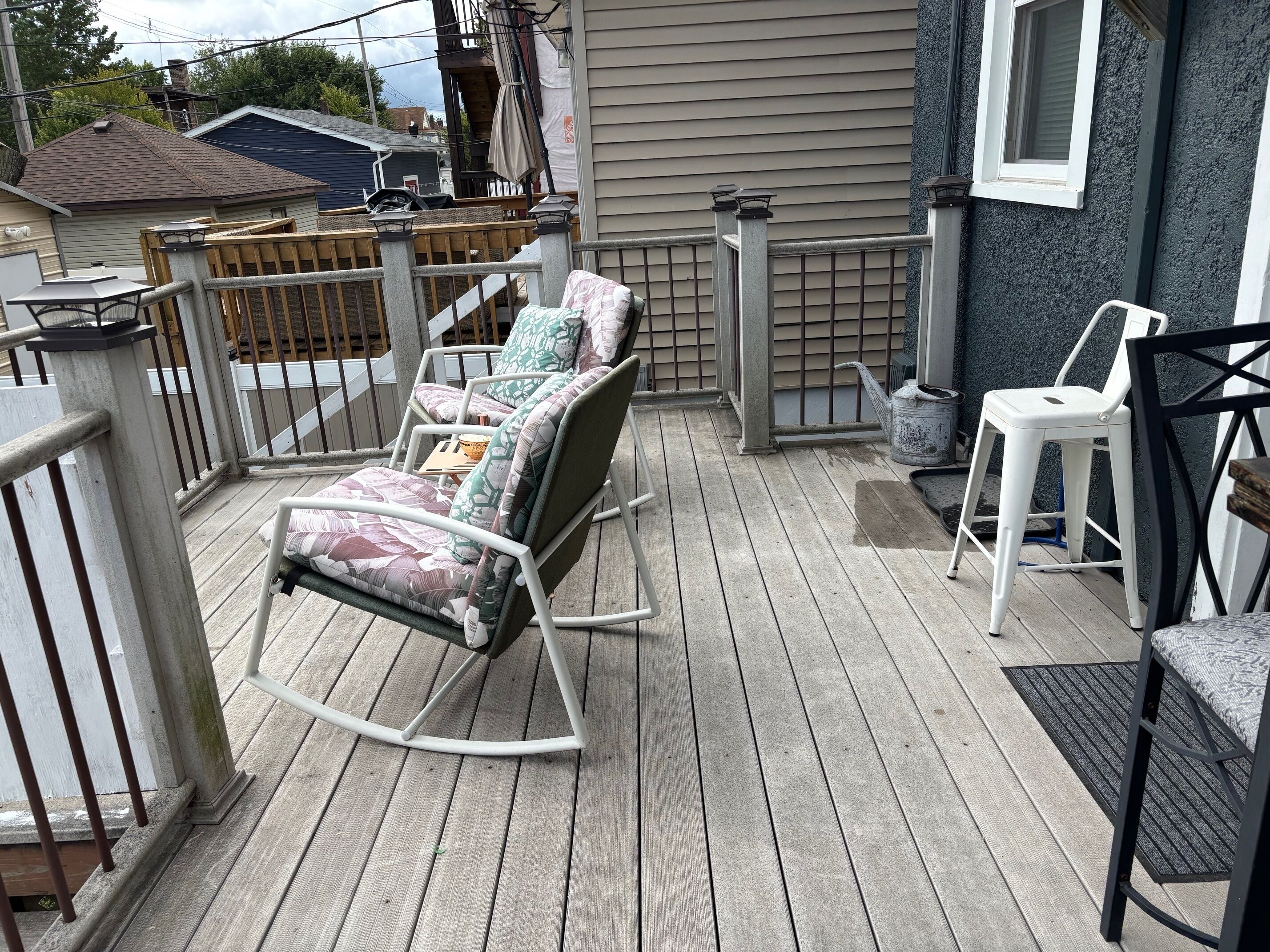 A wooden deck with a rocking chair and a white chair, a small table, and a metal railing.