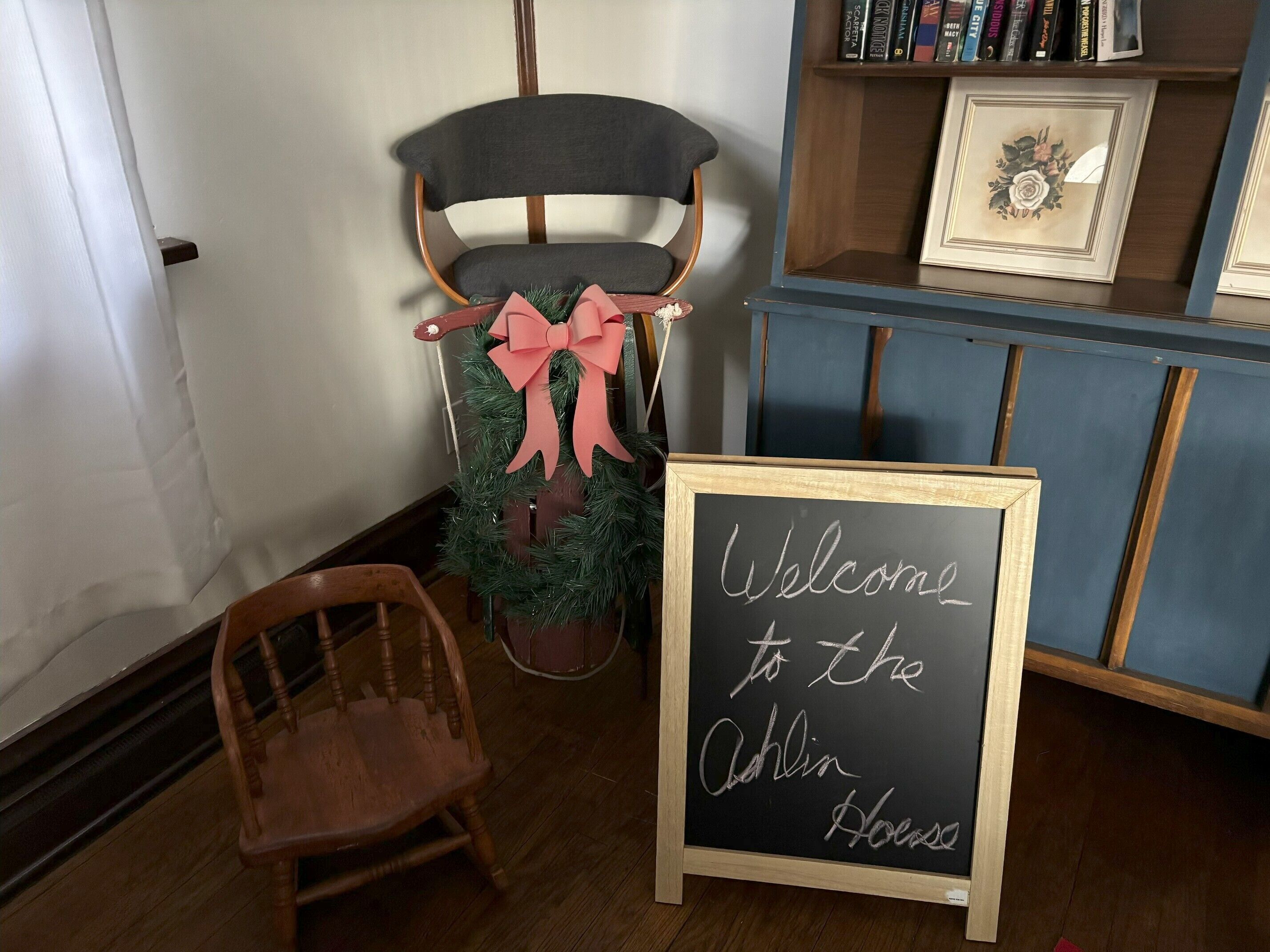 En træstol, et lille juletræ med et lyserødt bånd, og en tavle med teksten "Welcome to the Oaklin House".