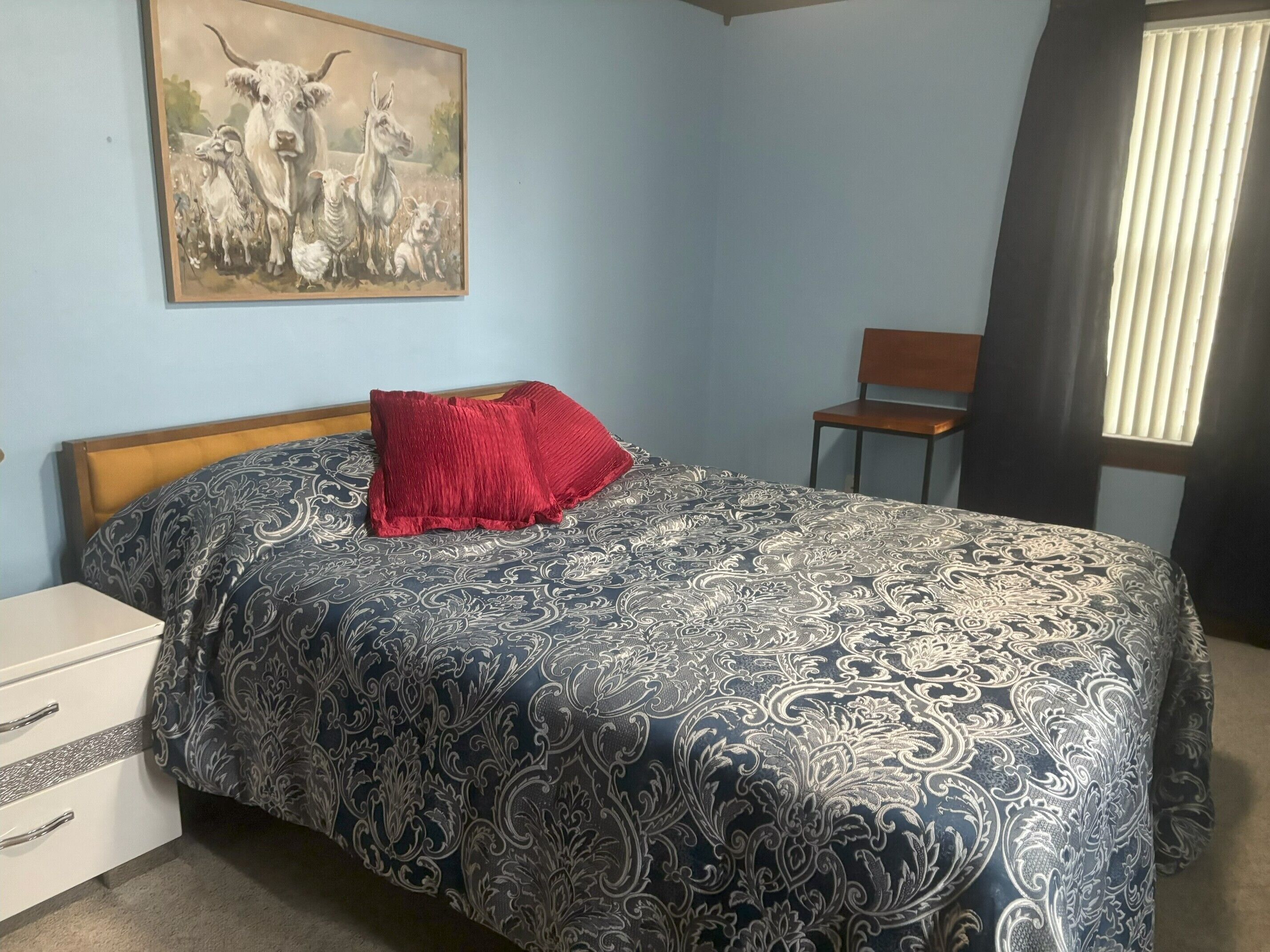 A bedroom with a bed, a nightstand, a chair, and a window with blinds.