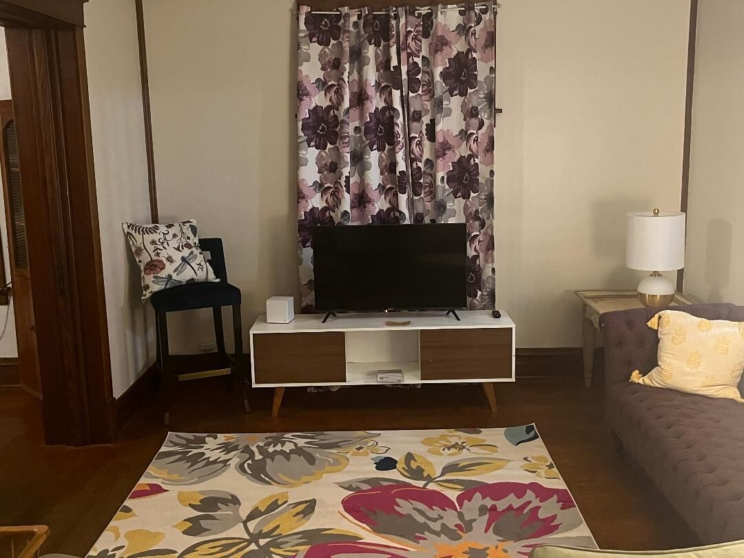 A living room with a flat-screen TV on a wooden TV stand, a floral-patterned rug, a purple sofa, and a small table with a lamp.